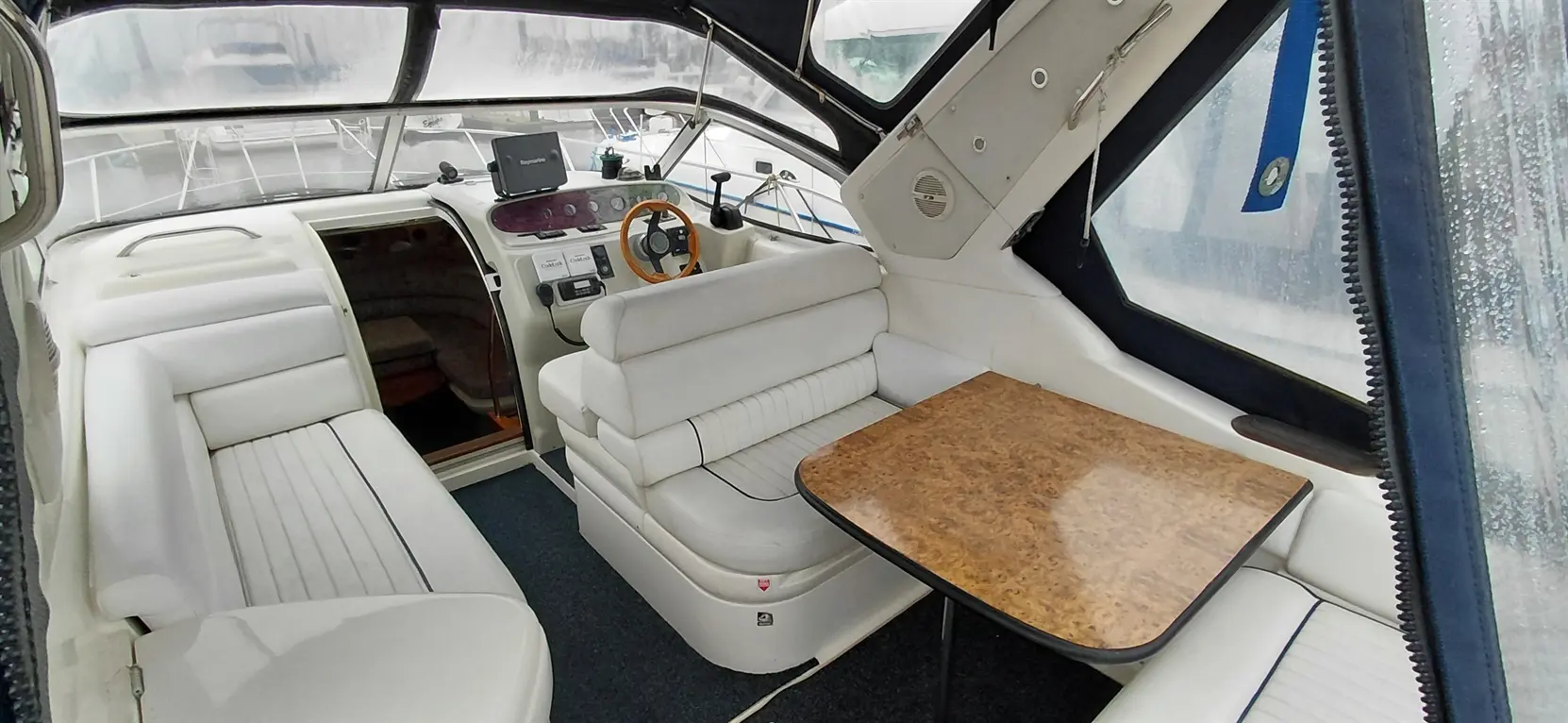 Interior view of a boat showing white seating, a wooden table, and a control panel with a screen, all in a modern design.