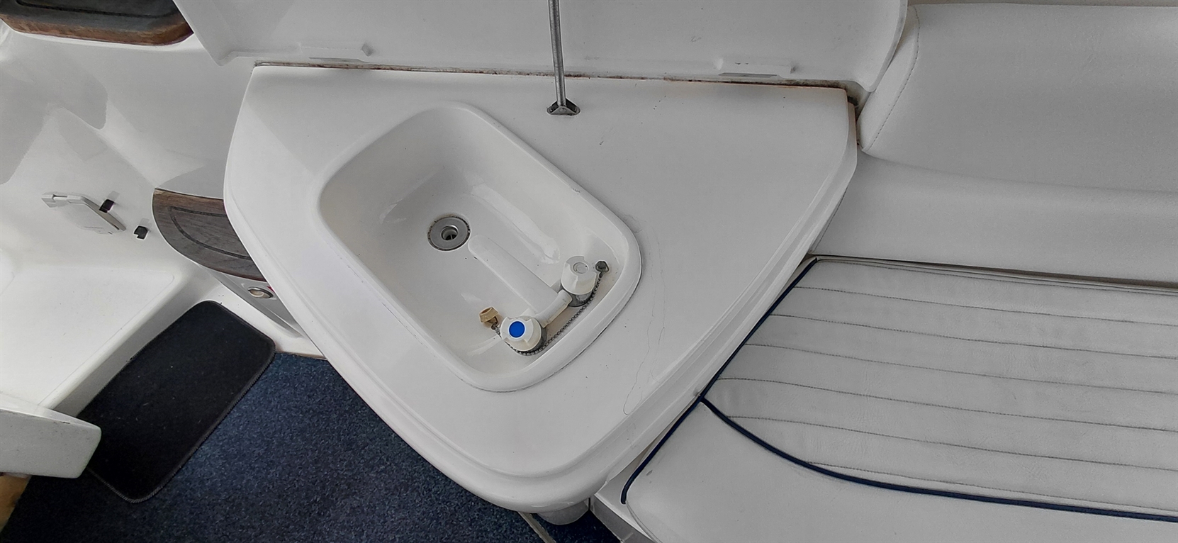White sink with faucet and plumbing fixtures on a boat, next to a white cushioned seat and dark blue carpet.