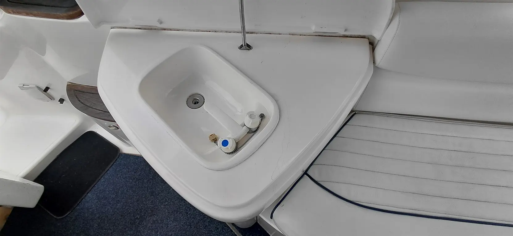 White sink with faucet and plumbing fixtures on a boat, next to a white cushioned seat and dark blue carpet.