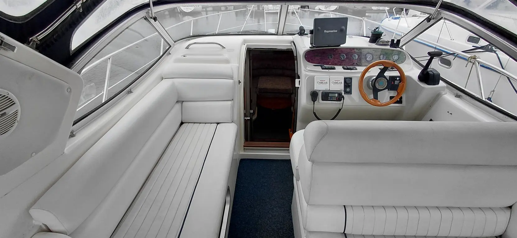 Interior view of a boat cabin showing white leather seating, dashboard controls, and a doorway leading to a cozy space.