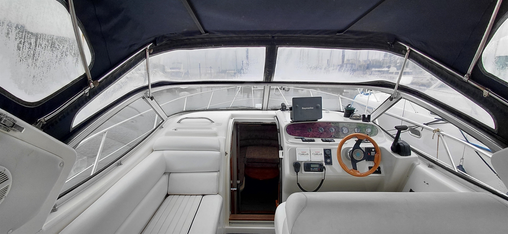 Interior of a boat showing white seating, steering wheel, and a dashboard with controls, overlooking a marina through foggy windows.