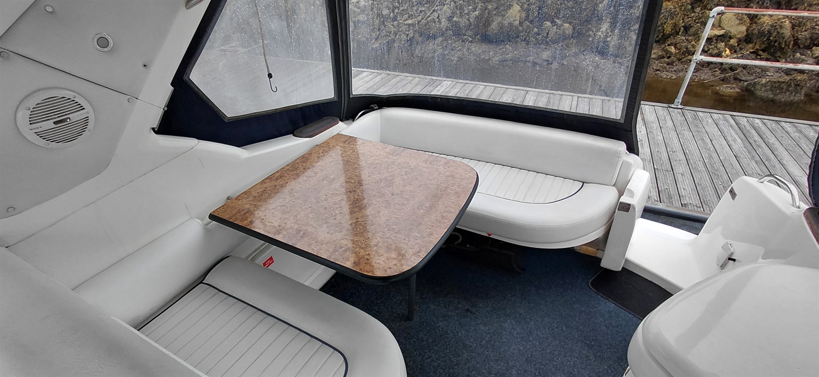 Interior of a boat featuring a wooden table, white seating, and a view of a wet dock through transparent windows.