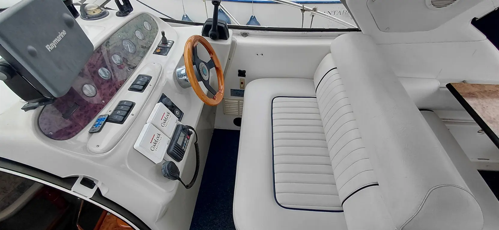 Boat cockpit view featuring a wooden steering wheel, control panel, and a white padded seat. Raymarine device visible.