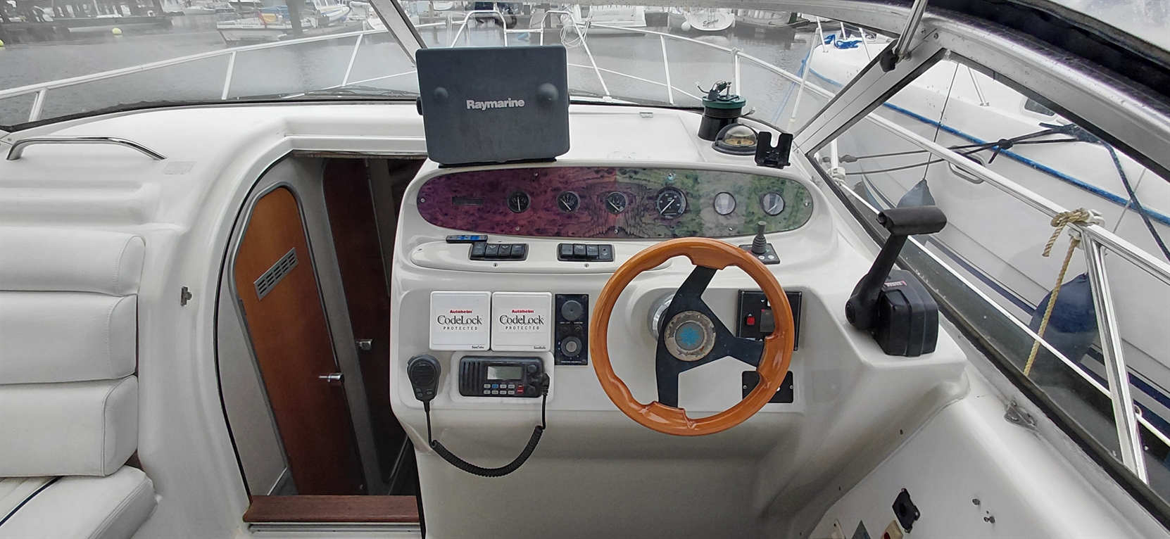 Boat cockpit featuring a wooden steering wheel, gauges, radio, and a Raymarine display, with a marina view through the windows.