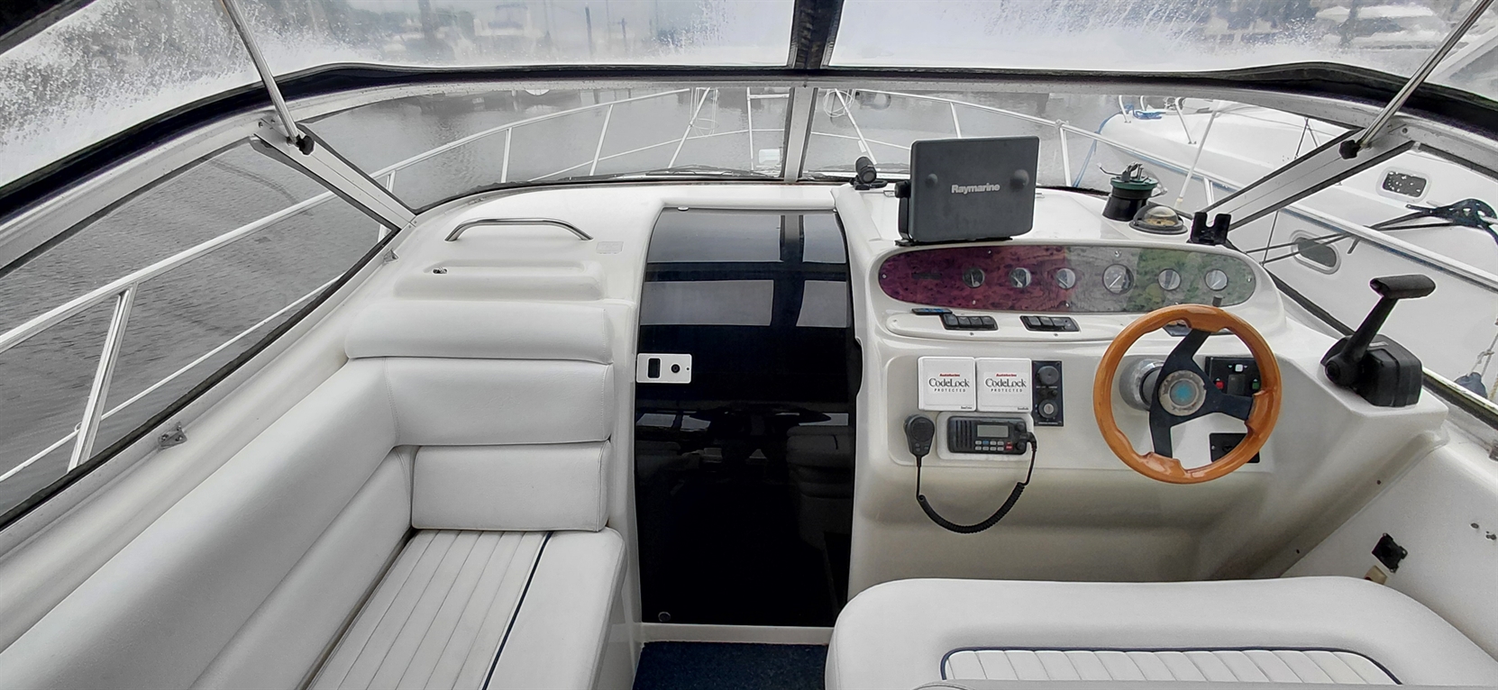 Boat interior featuring a steering wheel, control panel, and soft white seating, with a view of the water through windows.