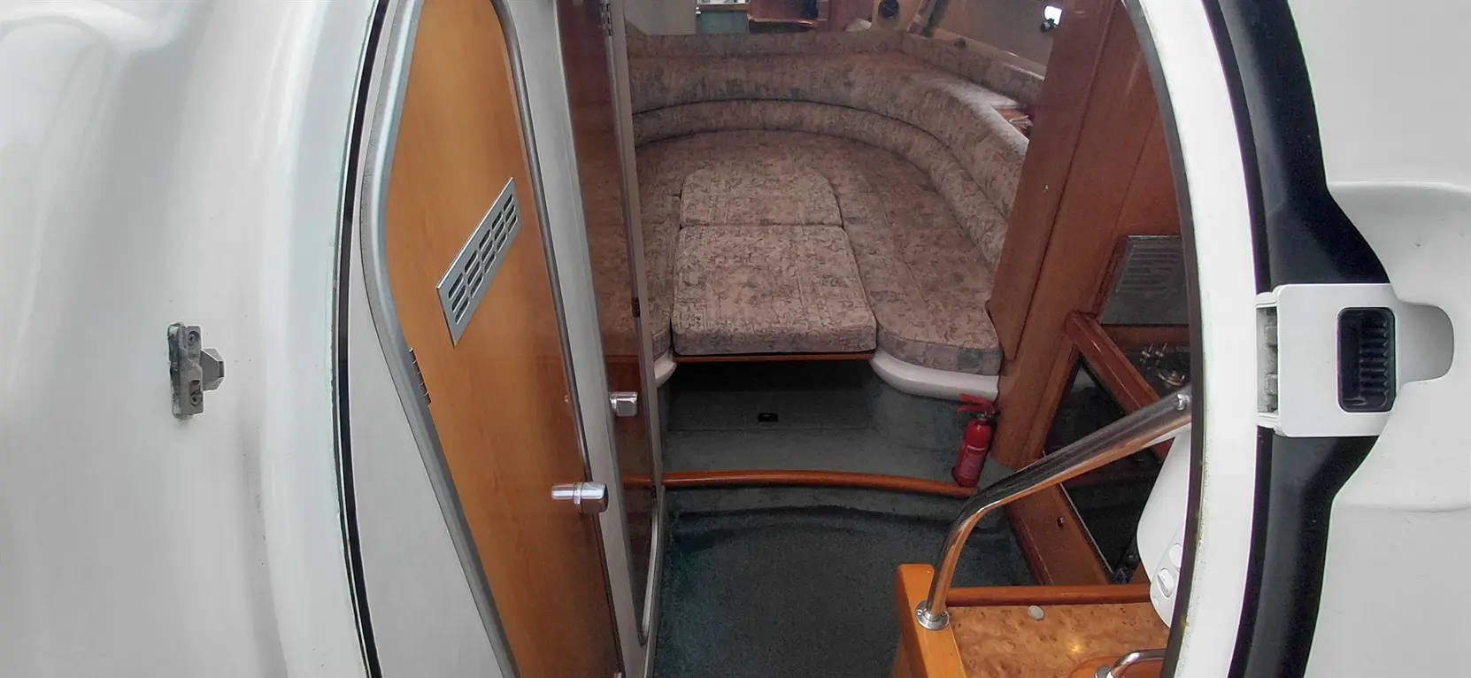 Interior view of a boat showing a wooden door, rounded seating with patterned upholstery, and fire extinguisher on the floor.