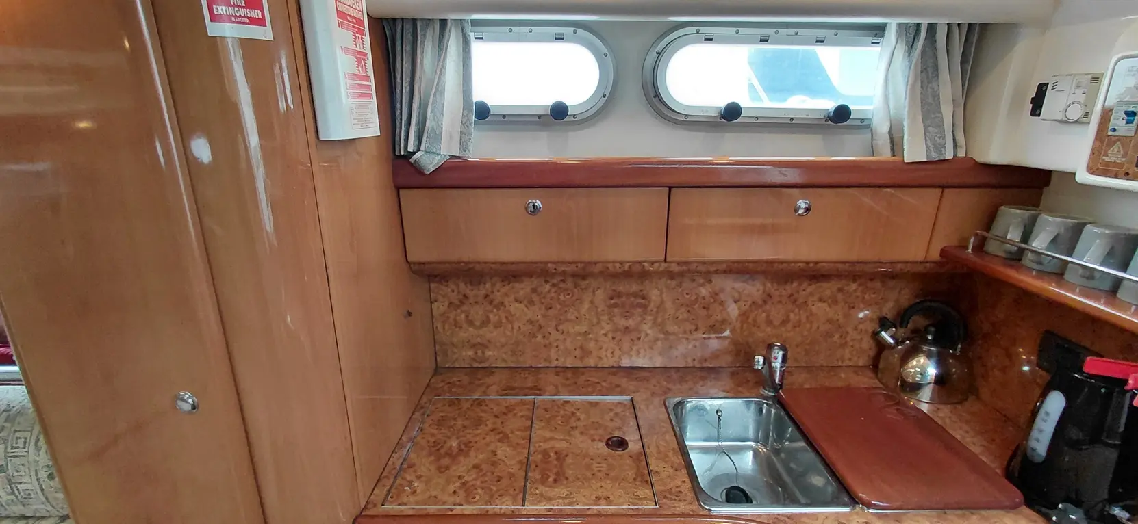 Wooden kitchen area in a boat, featuring a small sink, countertop, kettle, and storage drawers, with a fire extinguisher visible.