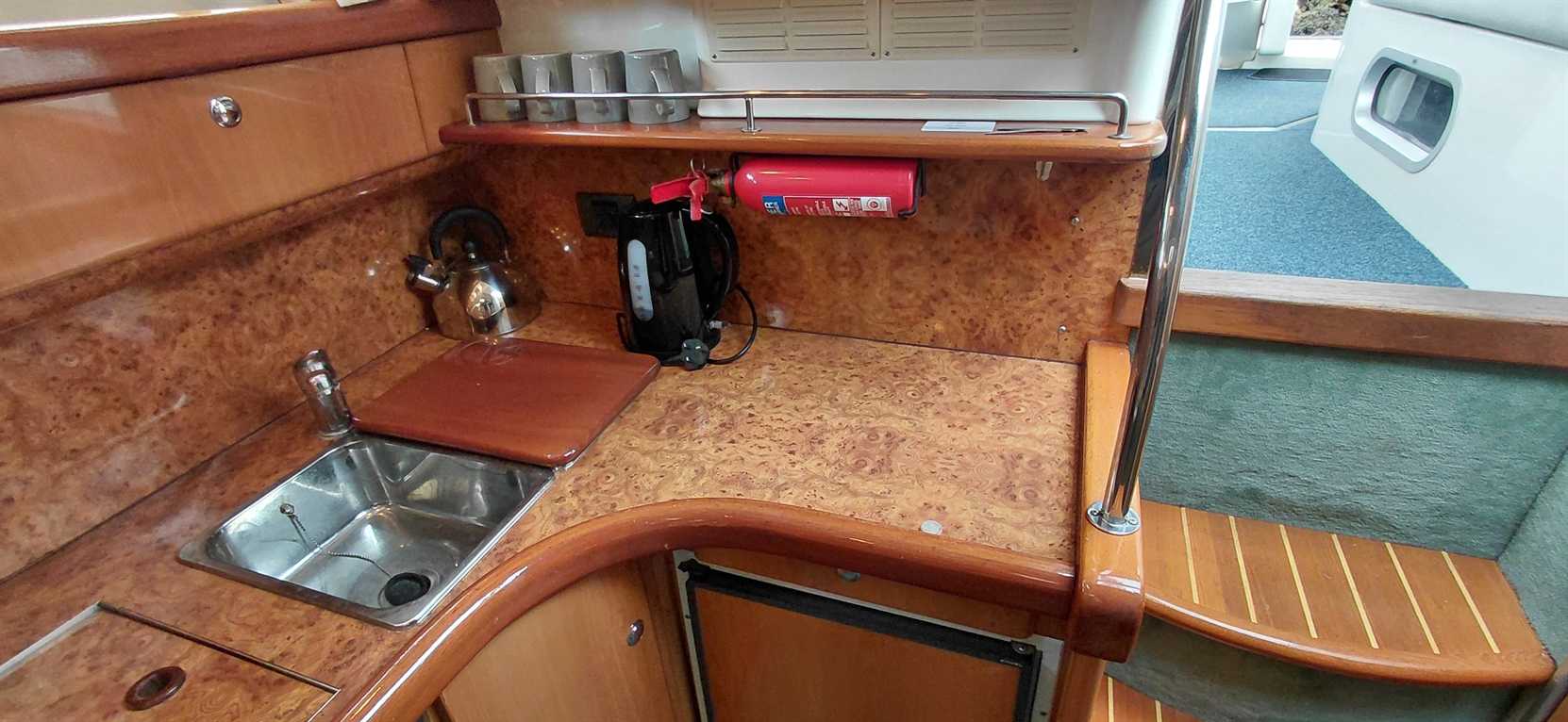 A compact boat kitchenette featuring a sink, kettle, fire extinguisher, and wooden countertop with mugs on display.