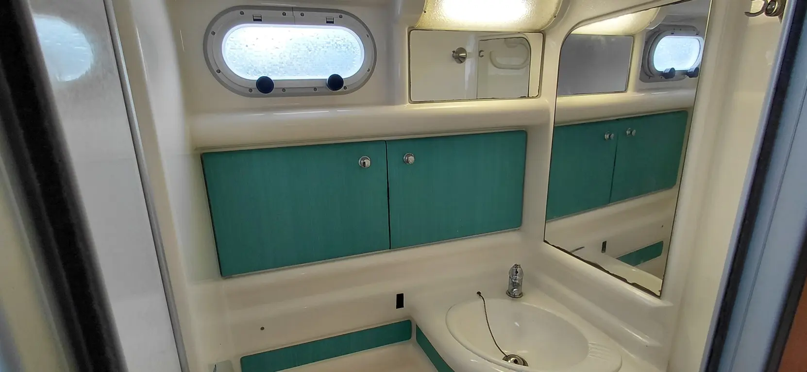 Compact bathroom with turquoise storage units, a sink, and a mirror, featuring a circular window letting in light.