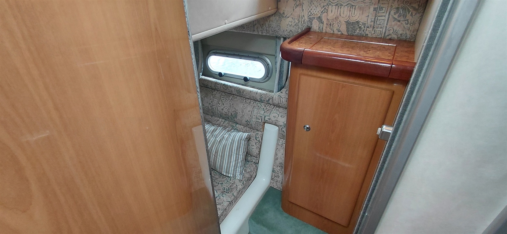 Interior of a small boat cabin featuring a striped cushion, wooden cabinets, and a round window. Soft green carpet visible.
