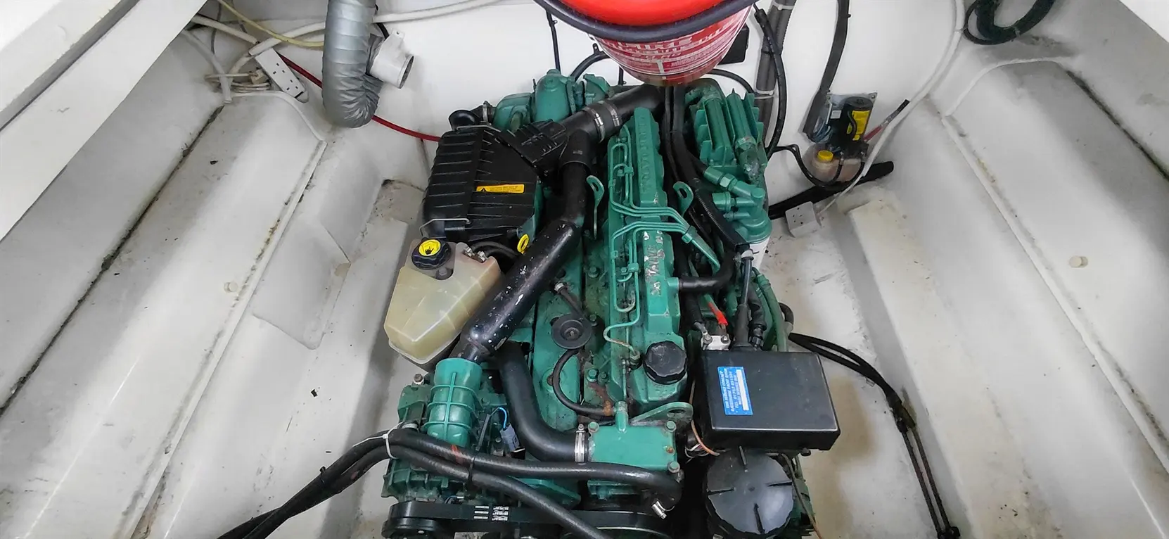 Boat engine compartment featuring a green marine engine with various hoses, a coolant reservoir, and a fire extinguisher above.