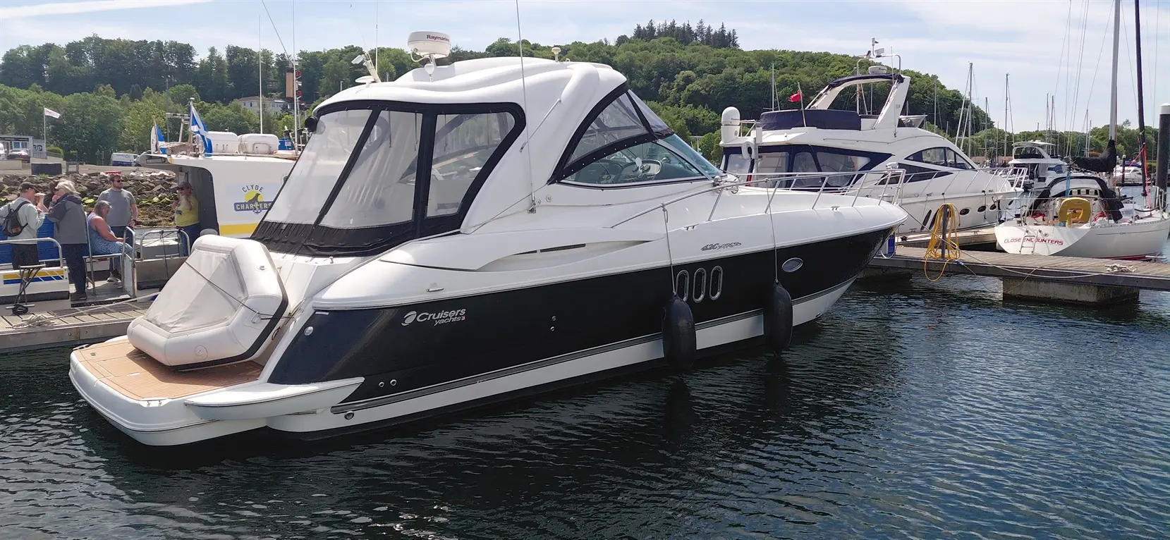 A sleek Cruisers Yachts boat docked at a marina, with people gathered on the adjacent dock enjoying the sunny day.