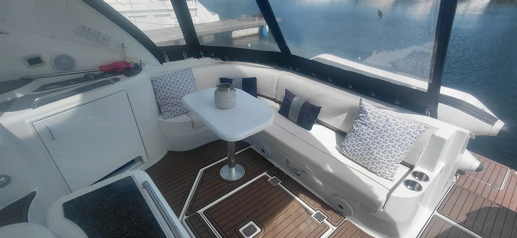 A modern boat interior featuring white seating, decorative pillows, a small table, and a view of the water outside.