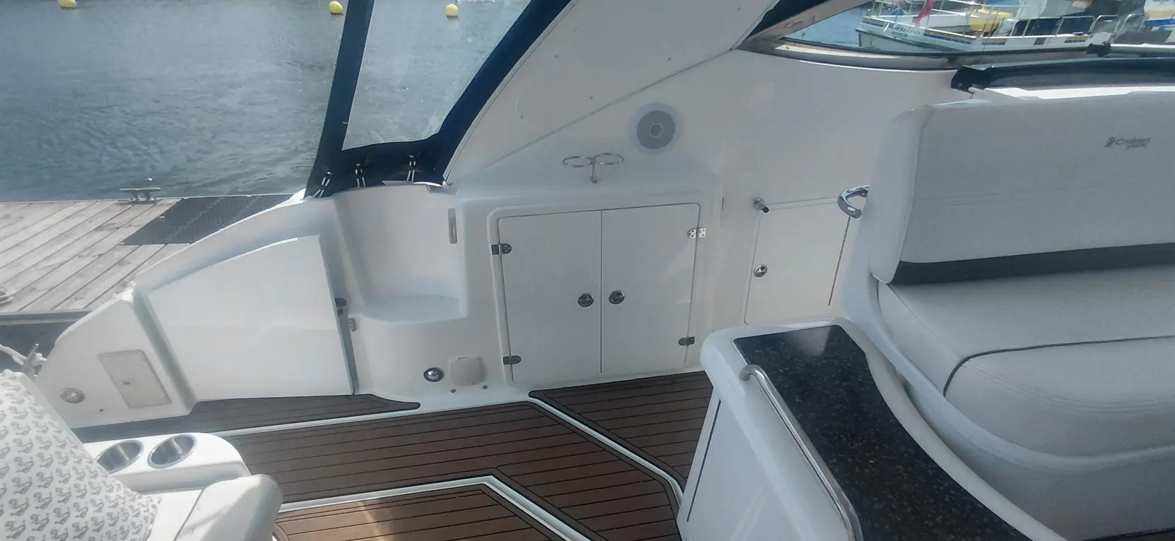 Interior view of a boat featuring a lounge seat, storage cabinet, and wooden flooring, with water and docks visible outside.
