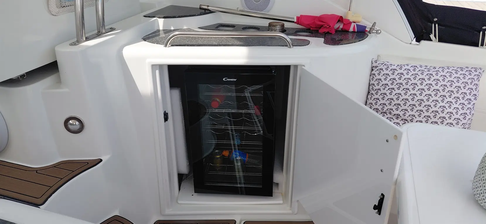 An open mini fridge in a boat's galley, filled with drinks, surrounded by a countertop and a patterned cushion.