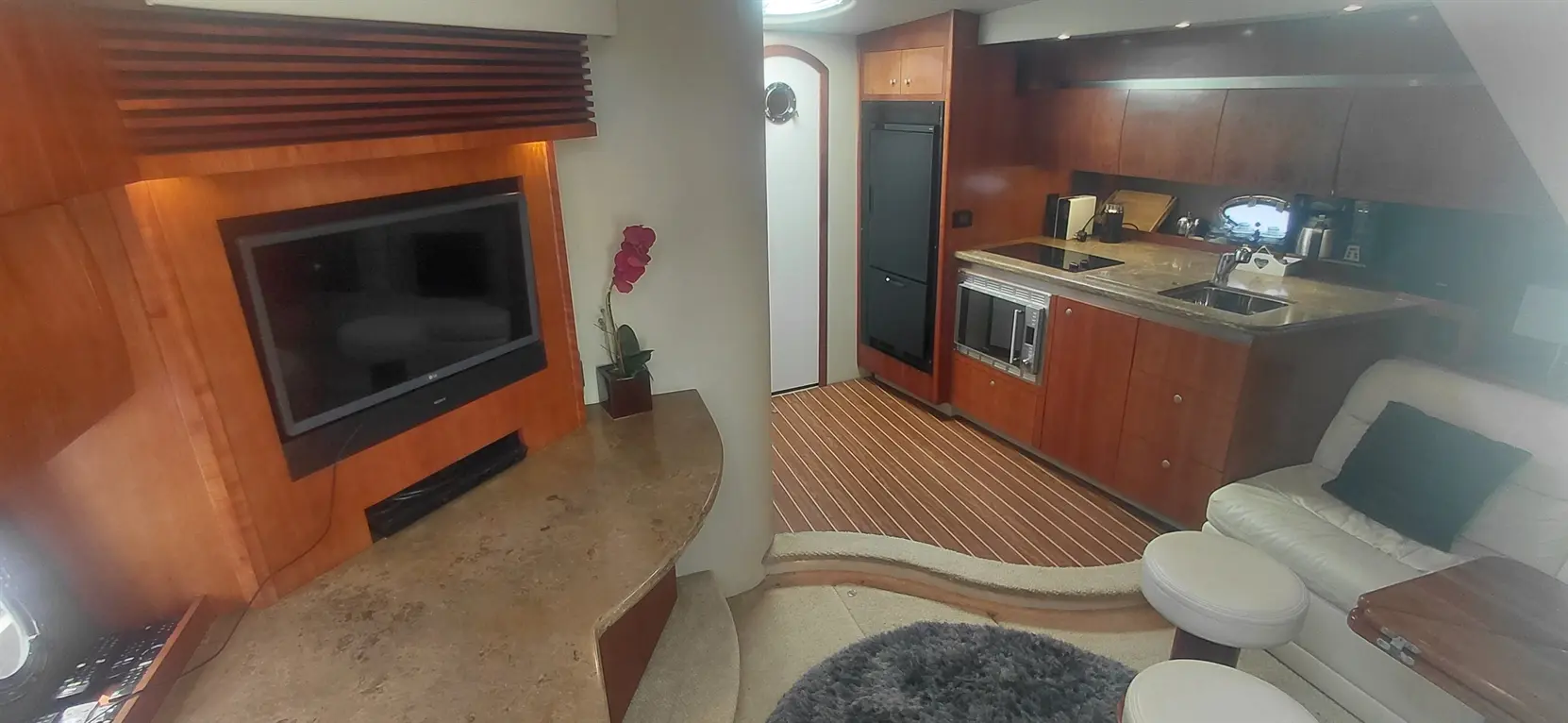 Modern yacht interior featuring a kitchen with wooden cabinets, a TV, and a cozy seating area with a rug and decorative orchid.