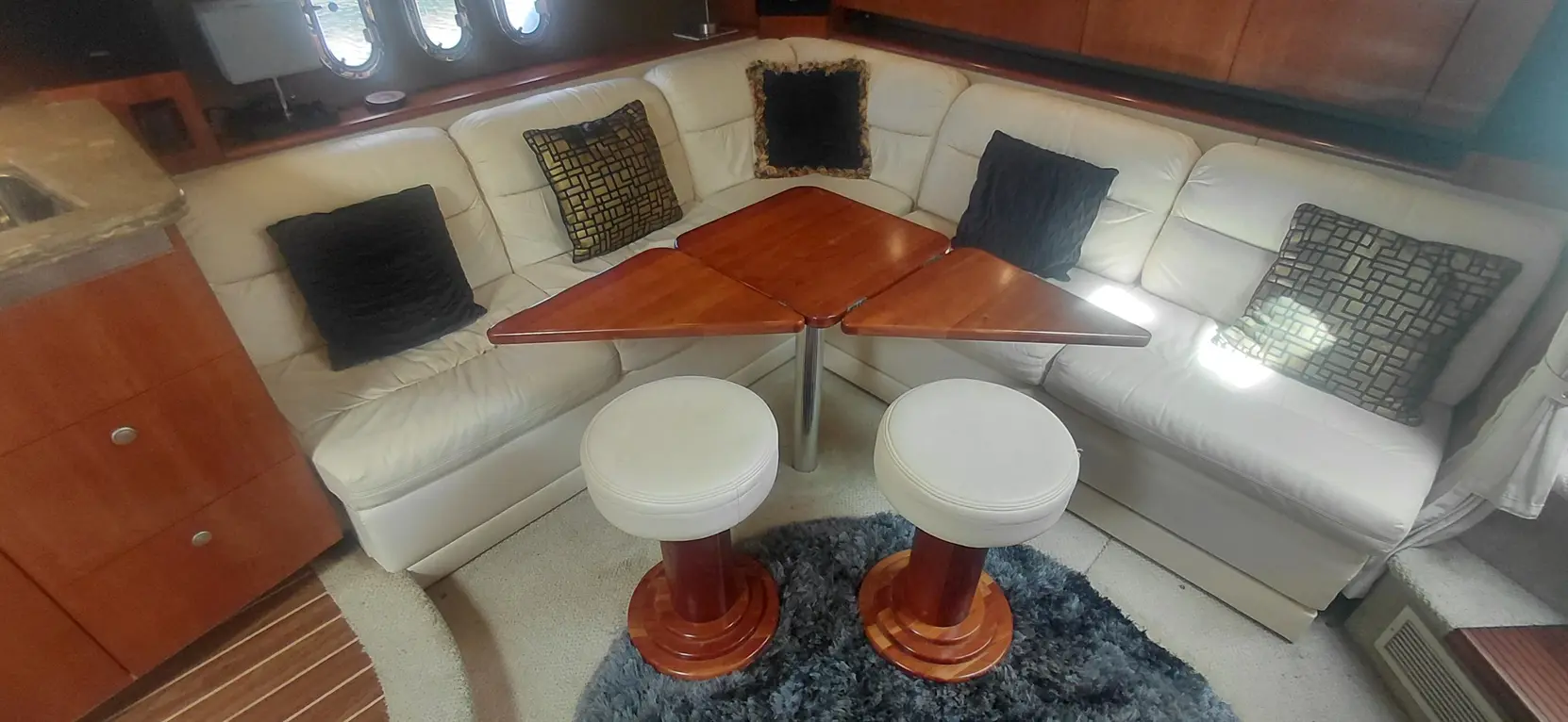 Cozy boat interior featuring a U-shaped white leather couch, triangular wooden table, and two white round stools on a textured rug.
