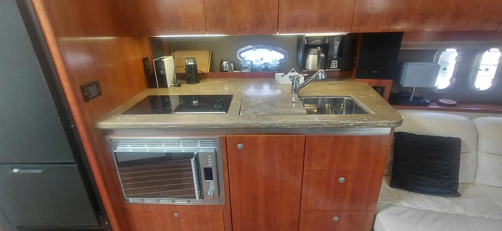 Modern yacht kitchen featuring a stone countertop, dual sink, cooktop, microwave, and wooden cabinetry with a couch nearby.