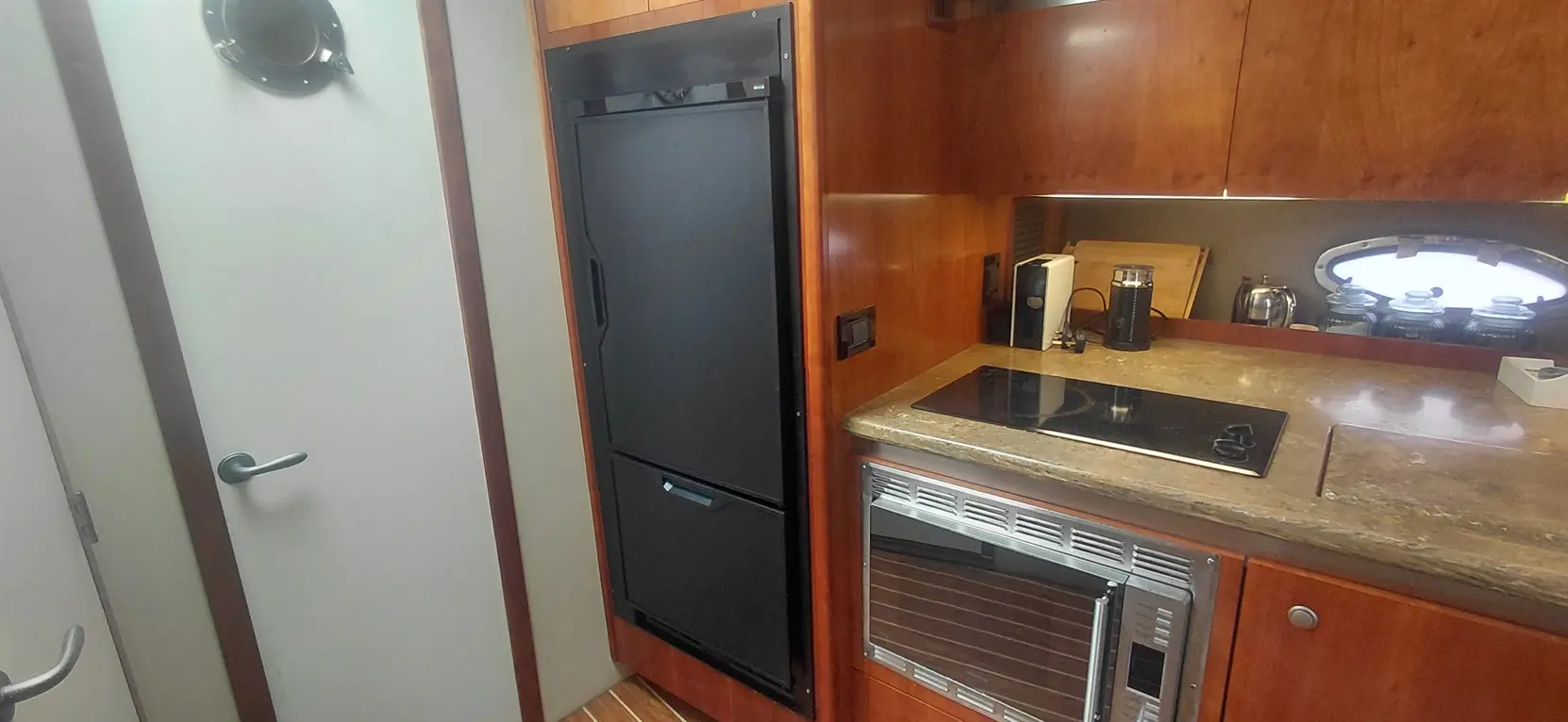 Modern boat kitchen featuring a black refrigerator, induction stovetop, and a microwave with wood cabinetry and a porthole window.