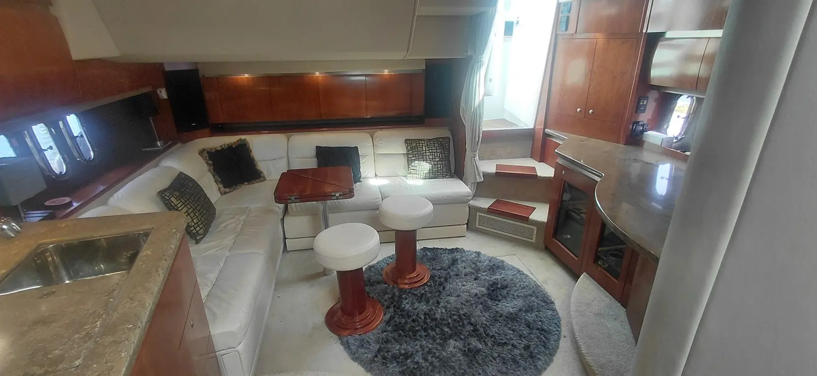 Luxurious yacht interior featuring a white sofa, round stools, a bar area, and polished wood accents, with windows offering natural light.