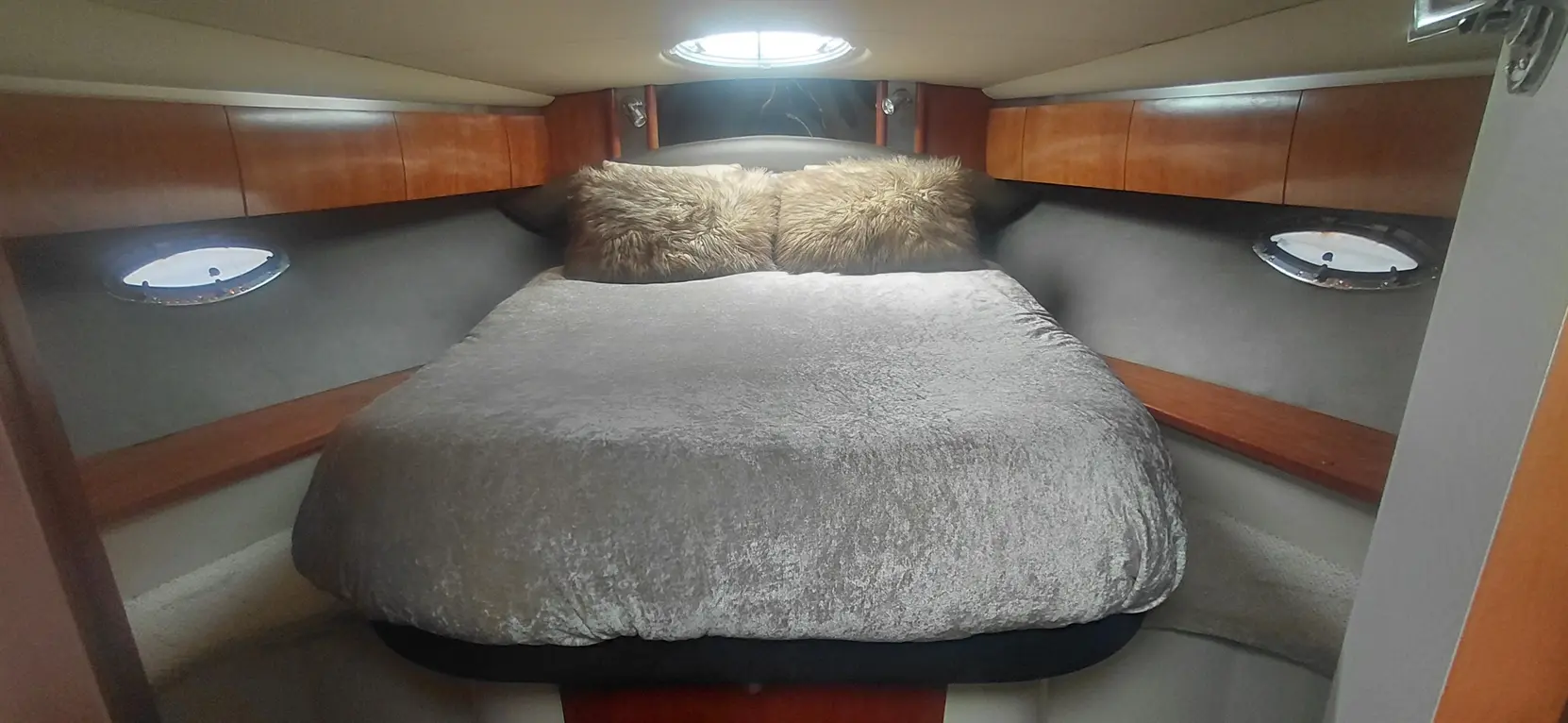 Cozy boat cabin featuring a double bed with a silver blanket and fluffy pillows, wood accents, and round porthole windows.