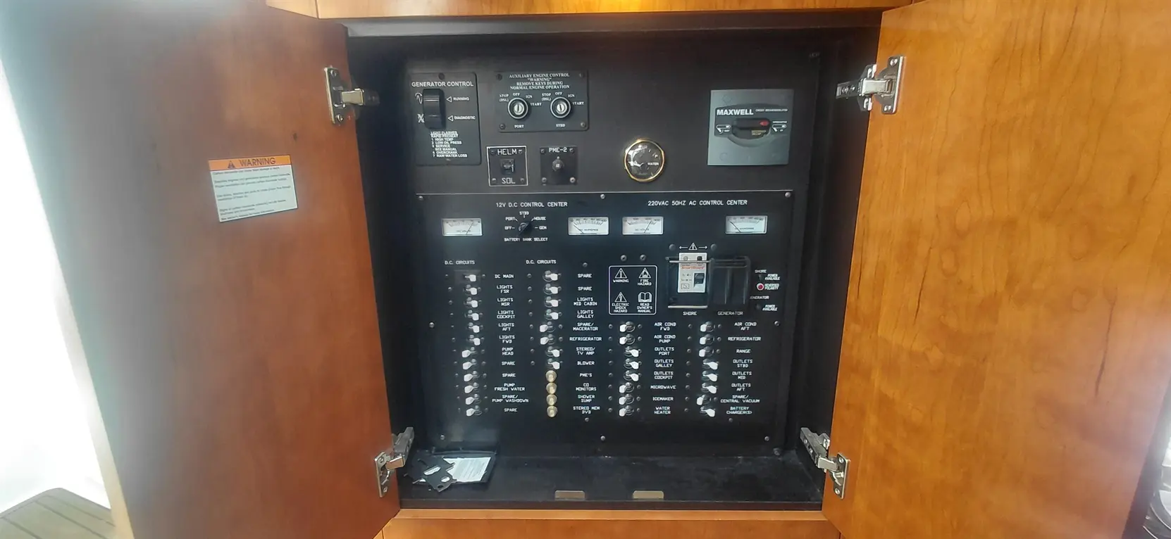 Panel with electrical controls, including generator and AC/DC systems, inside a wooden cabinet. Warning label visible.