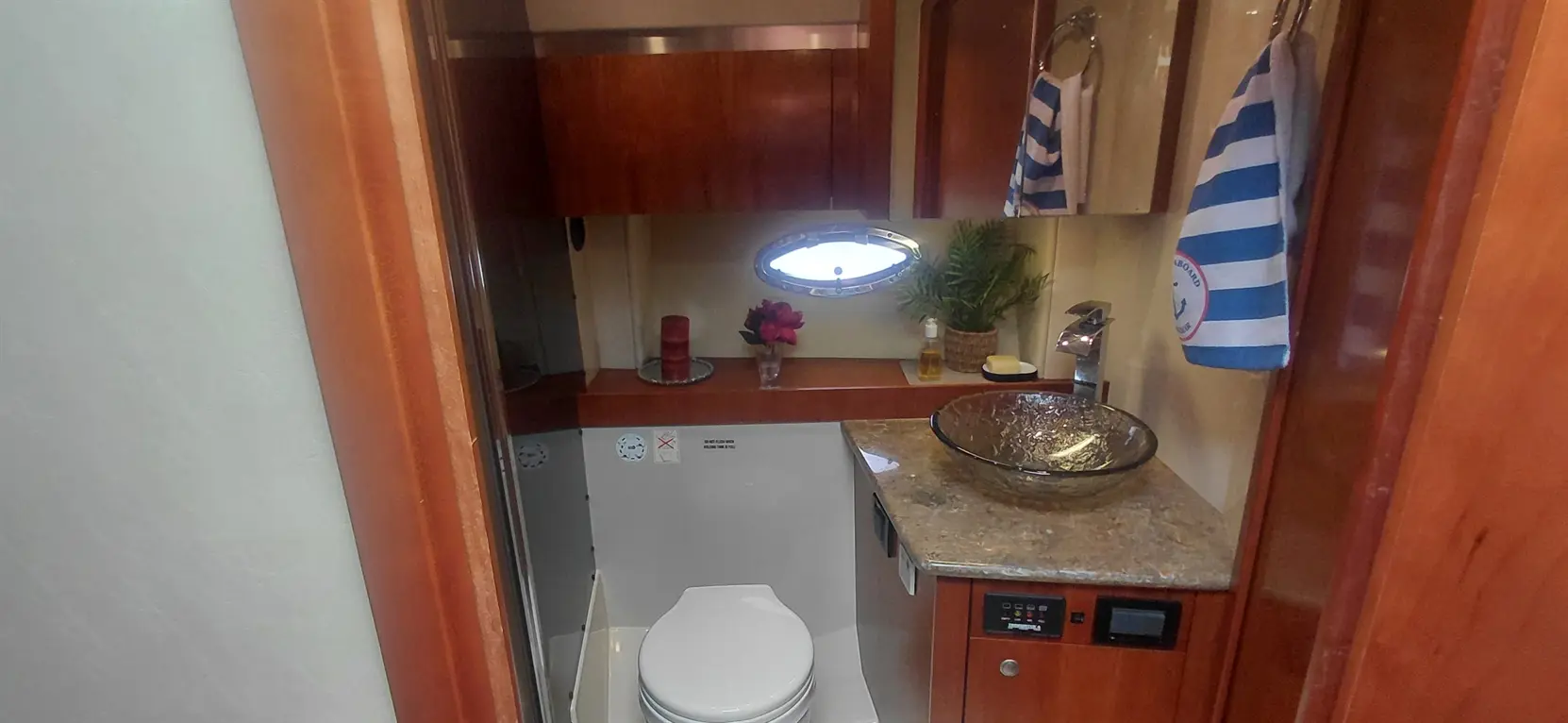 Modern boat bathroom with a white toilet, glass sink, decorative flowers, striped towels, and wooden cabinetry.