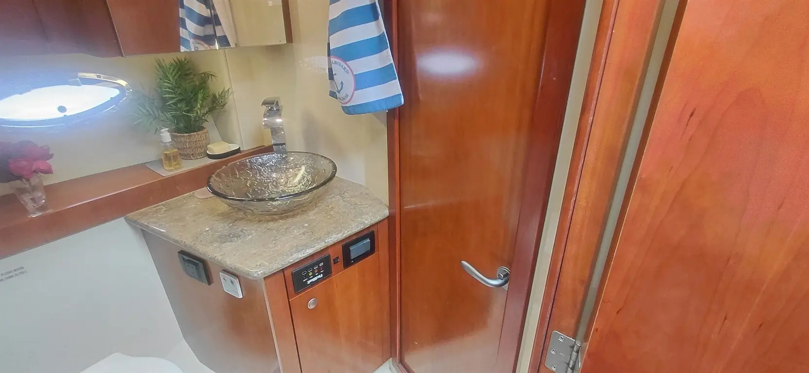 A modern boat bathroom featuring a glass sink, wood cabinetry, and a towel hanging, with decorative plants and floral arrangements.