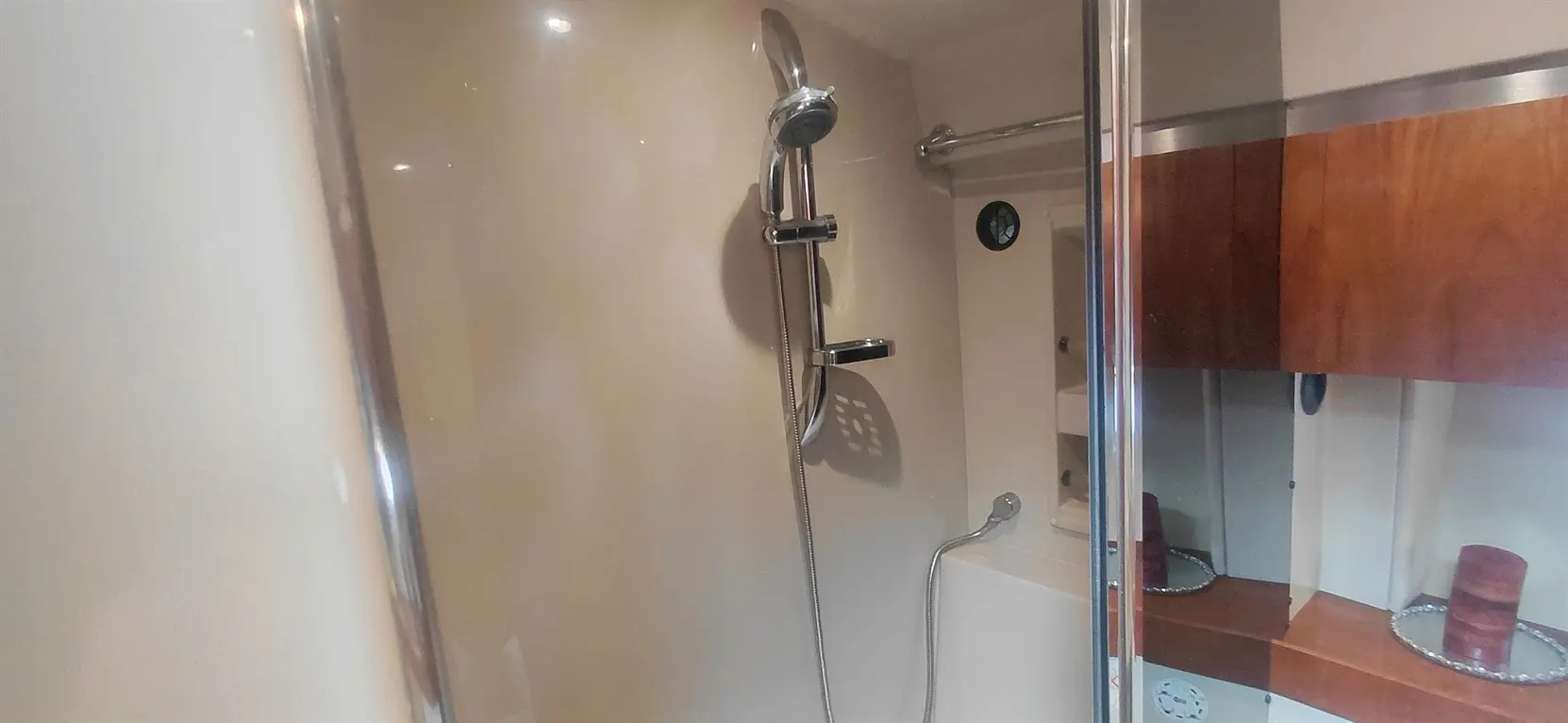 A modern shower stall featuring a chrome showerhead, glass door, and light beige walls, with wooden shelves in the background.
