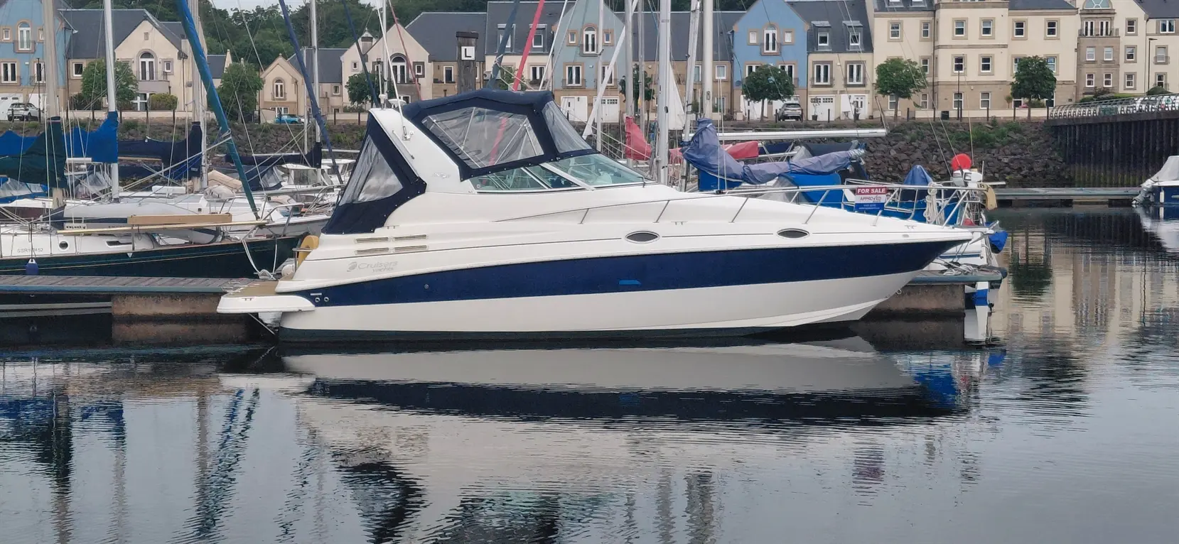 A cruiser boat docked at a marina, reflecting in the water, surrounded by other boats and waterfront buildings.