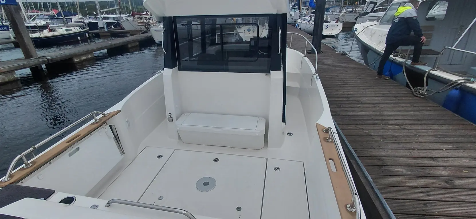 Empty boat deck with a seating area, near a dock filled with yachts and a person sitting on a wooden deck.
