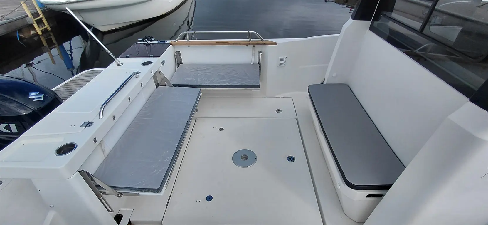View of a boat's deck with gray cushions, a mounted railing, and a backup motor visible in the background on calm water.
