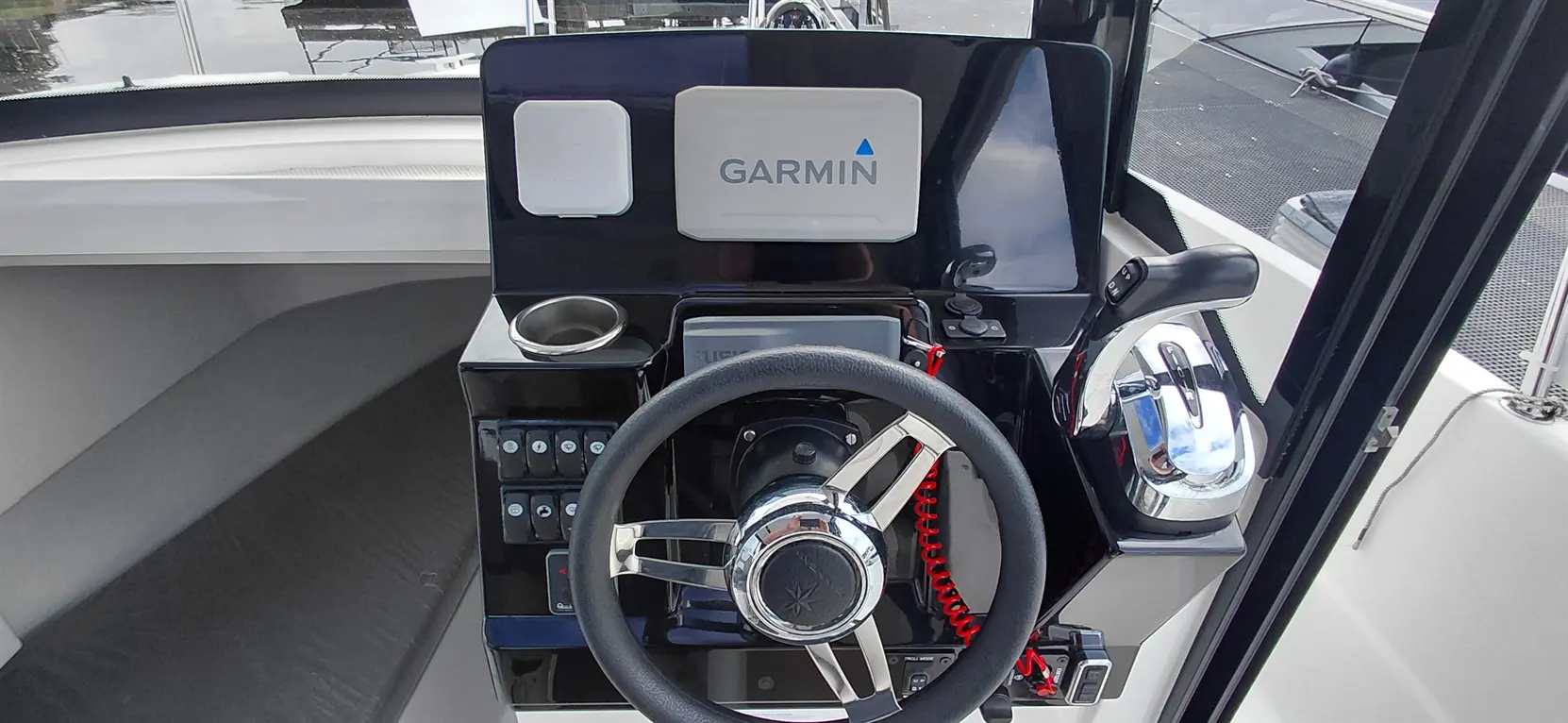 A boat's cockpit featuring a Garmin display, steering wheel, control knobs, and a cup holder with a serene water background.