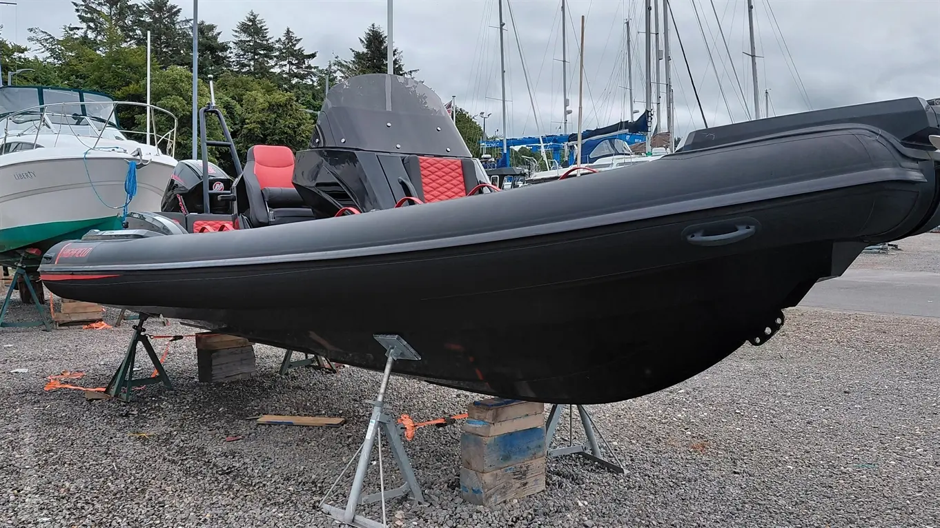 A black rib boat with red accents, supported by stands on gravel, surrounded by other boats and a cloudy sky.