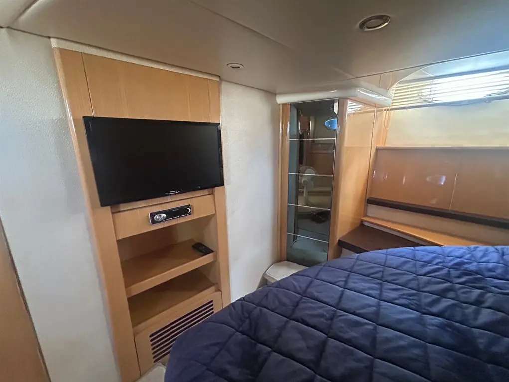 Cozy boat interior featuring a wall-mounted TV, shelf, and a double bed with a blue quilted cover. Natural light from a window.