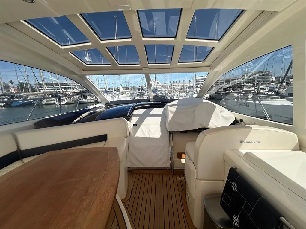 Interior view of a yacht cabin with large windows, wooden table, and visible boats docked outside. "ABSOLUTE" logo on chair.