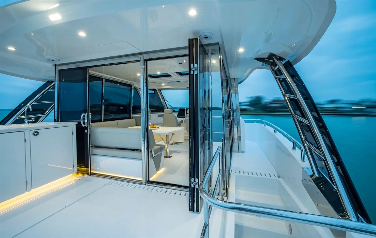 Modern yacht interior showing sleek white finishes, spacious seating area, glass doors, and views of water at dusk.