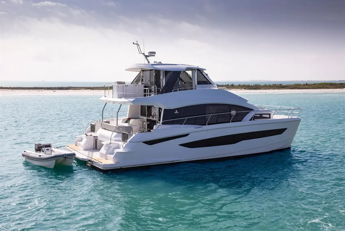 Luxury motor yacht anchored in clear turquoise waters, with a small tender boat alongside and a sandy beach in the background.