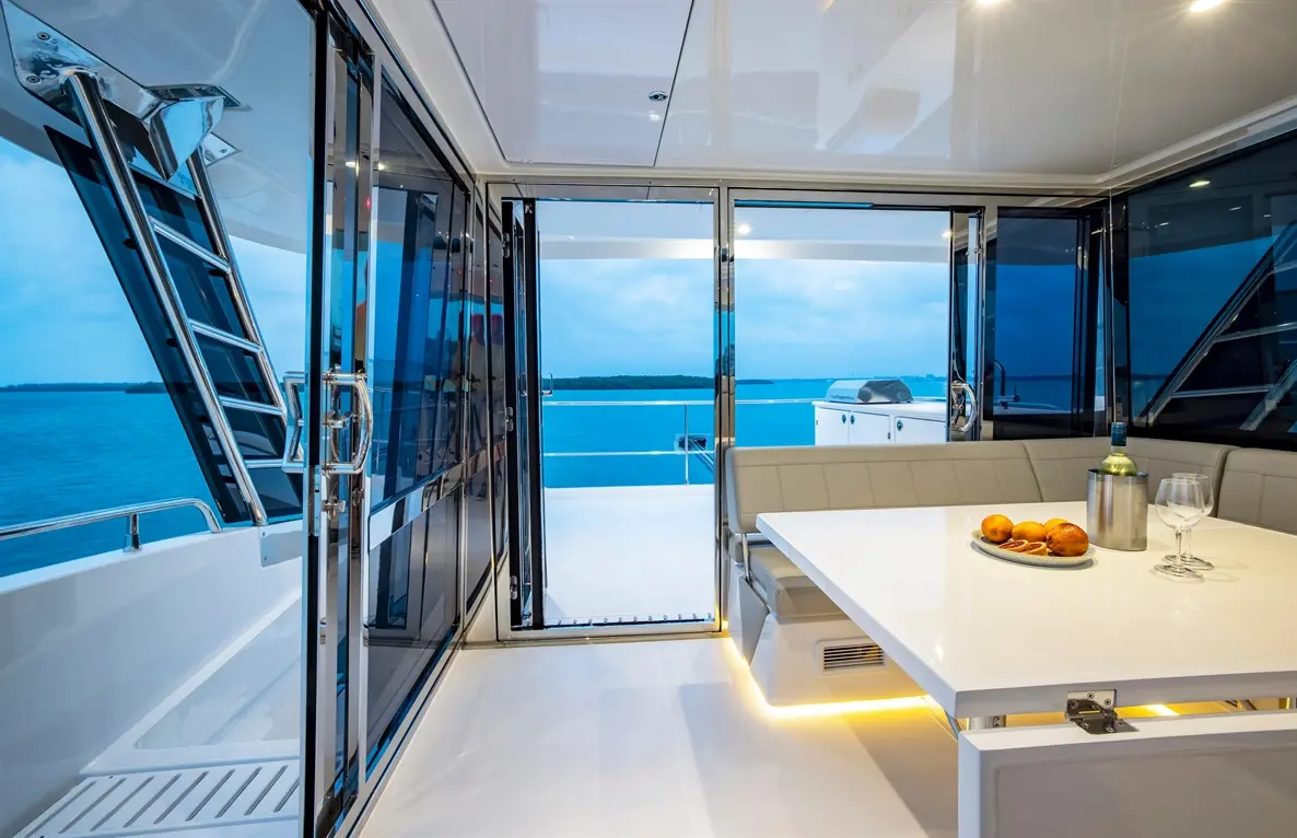 Modern yacht interior with large glass doors revealing a serene blue sea, a dining table set with fruits and a drink.