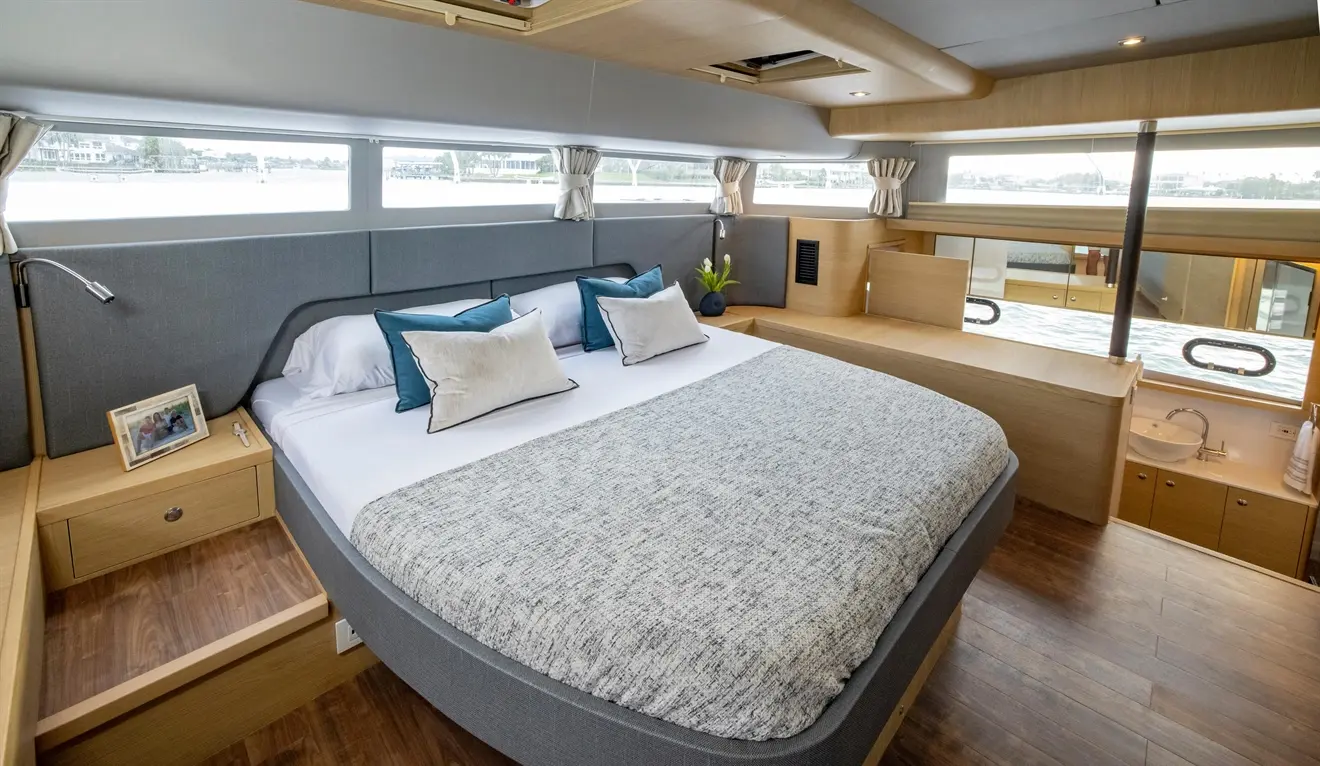 Stylish boat bedroom featuring a large bed with grey and teal cushions, wooden furnishings, and scenic water views through windows.