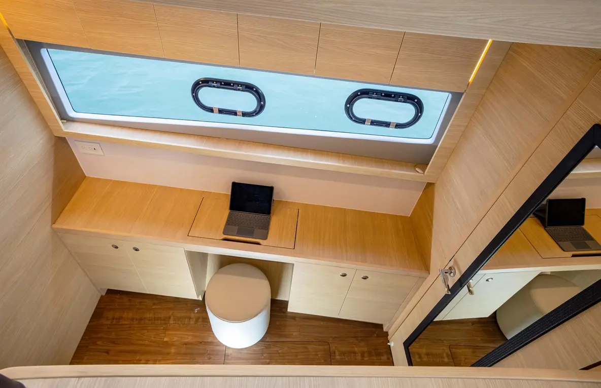 Modern office space on a boat features a desk with a laptop, round stool, and wide window showing tranquil water views.