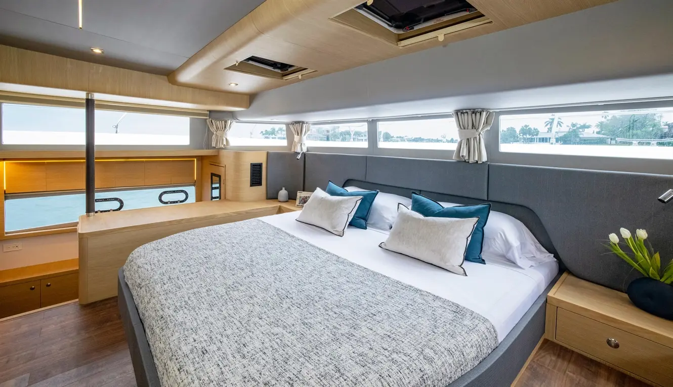 Modern boat bedroom with large windows, grey and blue décor, a double bed, and a wooden dresser. Beautiful water view outside.