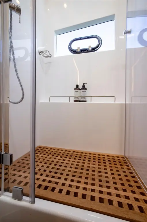 Modern shower with wooden slatted floor, metal showerhead, and bottles of shower gel on a shelf. Natural light enters through a window.