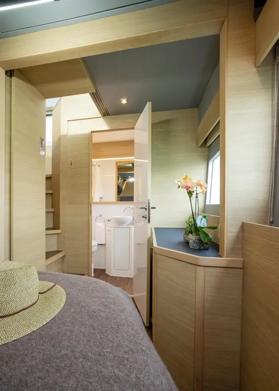 A modern wooden interior of a cabin, featuring a bed with a hat, a bathroom door, and a potted orchid on the counter.