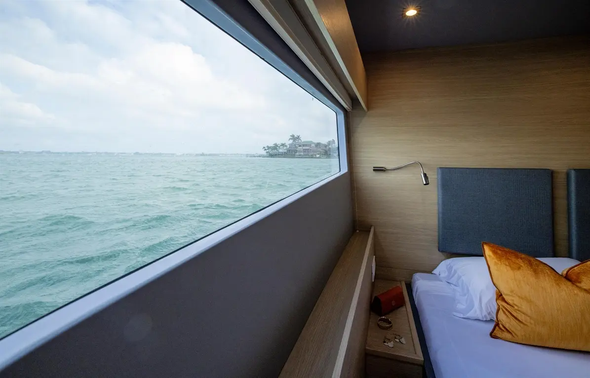 Modern bedroom with a large window overlooking the calm sea and distant shore, featuring a bed and decorative cushions.