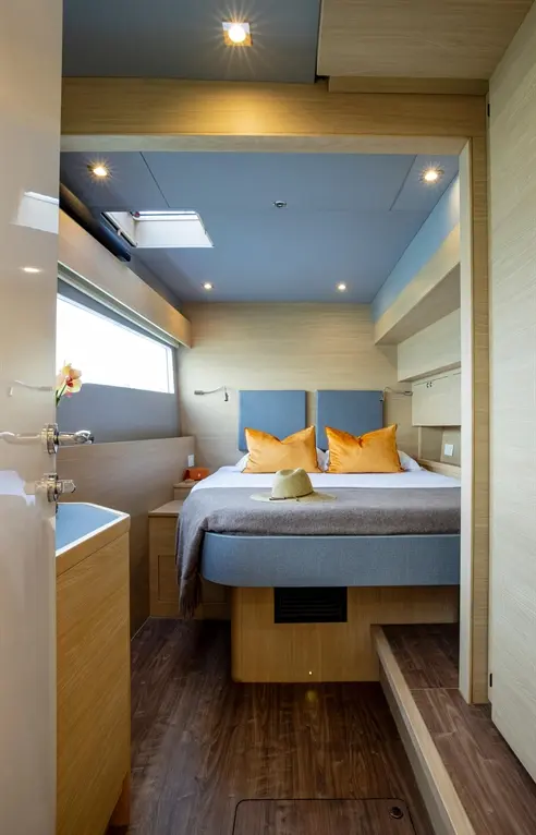 Modern yacht cabin featuring a double bed with orange cushions, a straw hat, and a window for natural light.