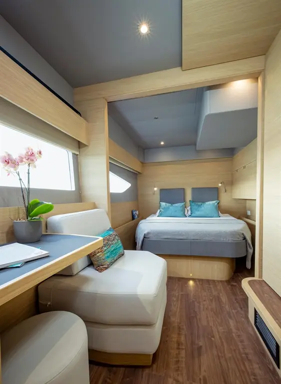 Modern yacht interior featuring a bed, seating area, desk, and potted orchids, with light wooden finishes and large windows.
