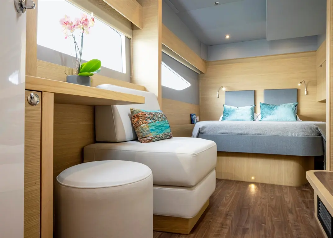 Modern interior of a cabin with a double bed, armchair, ottoman, and decorative orchids by a window, featuring wood finishes.