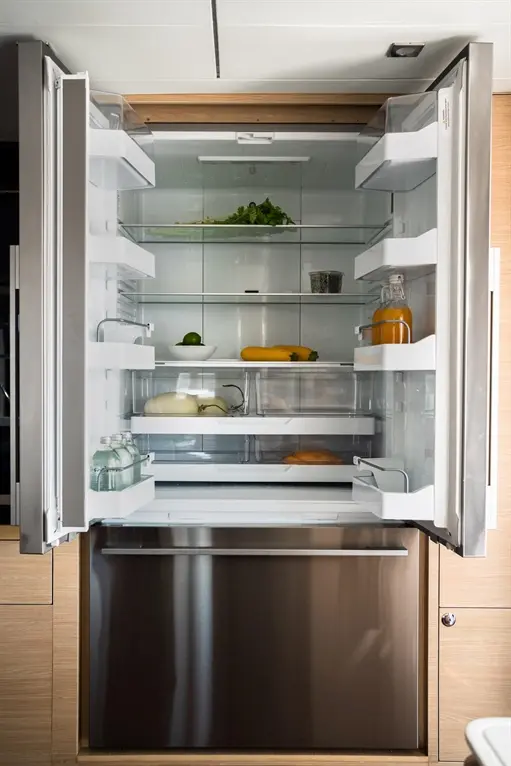 An open modern fridge with glass shelves, stocked with fresh vegetables, drinks, and dairy products, against a wooden background.