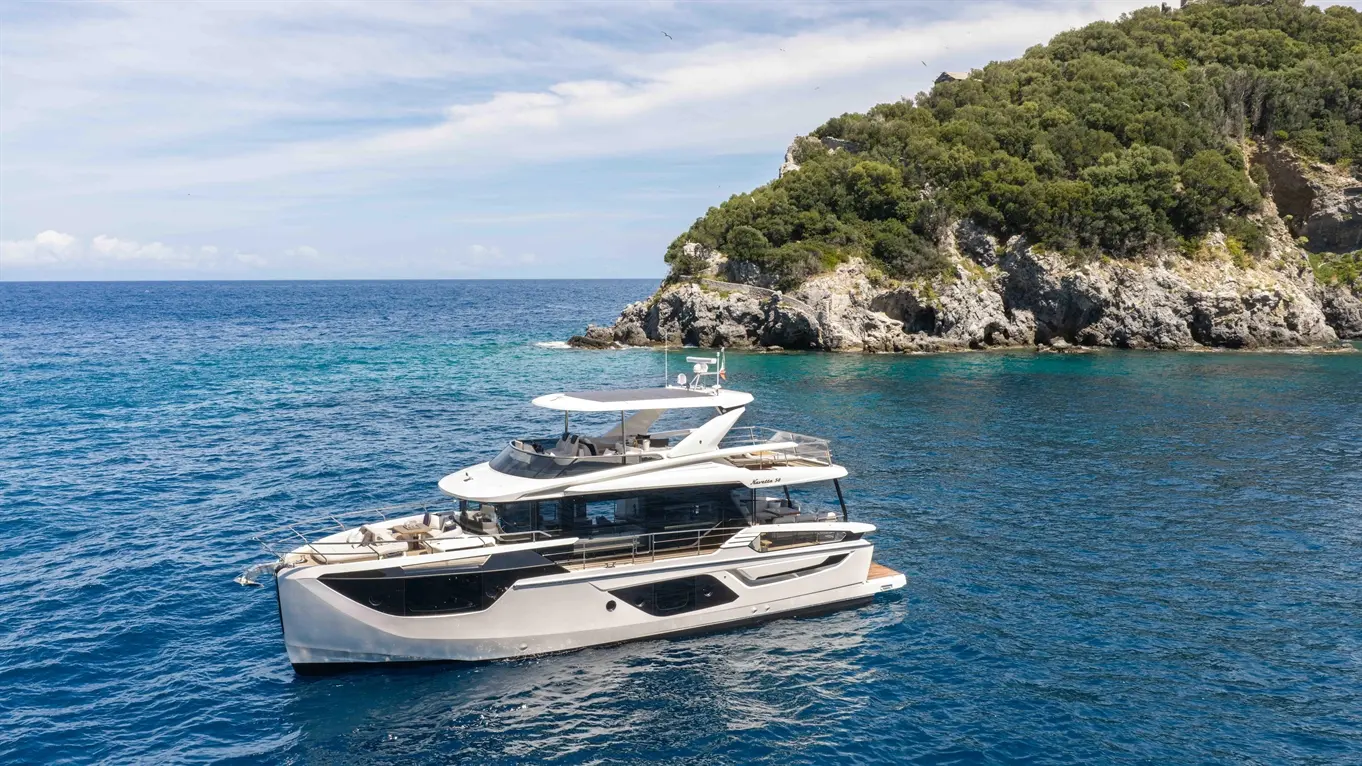 A luxury yacht anchored by a rocky coastline, surrounded by clear blue waters and lush greenery under a partly cloudy sky.